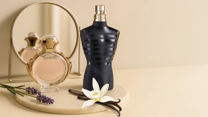 Zwei Parfums auf heller Marmorplatte mit Spiegel im Hintergrund sowie Lavendel und Vanilleblüte im Vordergrund.